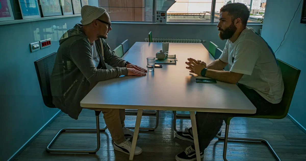 Dos hombres conversando frente a frente en una mesa, junto a una ventana.