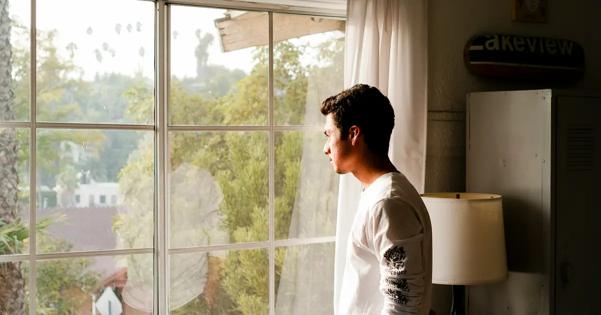 Hombre joven de pie junto a una ventana, mirando hacia afuera en un momento de reflexión